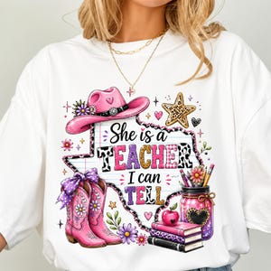 May include: A white t-shirt featuring a graphic of a pink cowboy hat, pink cowboy boots with purple bows, a leopard print star, and a mason jar filled with pencils and an apple on books. The text reads, She is a TEACHER I can TELL.