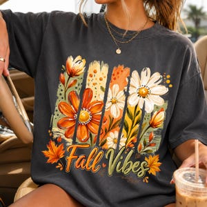 May include: A charcoal gray short-sleeve t-shirt featuring a graphic print of orange and white flowers, green leaves, and autumn maple leaves. The text "Fall Vibes" is printed in a cursive script at the bottom of the floral design.