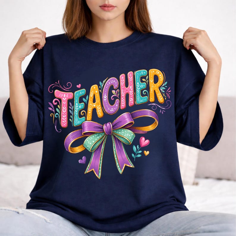 May include: A navy blue t-shirt featuring the word TEACHER in colorful, patterned block letters. Below the text is a large, decorative purple and teal bow with intricate floral details and small heart accents, creating a vibrant graphic design.