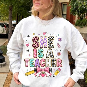 May include: A white long-sleeve sweatshirt featuring the text She Is A Teacher in colorful patterned letters. The design includes a yellow ruler, a pink and white plaid bow, a red apple, and a yellow pencil, surrounded by small hearts and flowers.