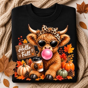 May include: A black sweatshirt featuring a graphic of a fluffy brown highland cow wearing leopard print sunglasses and a matching bow. The cow is blowing a pink bubble, holding a coffee cup, and standing near pumpkins, autumn leaves, and a sign reading Hello Fall.