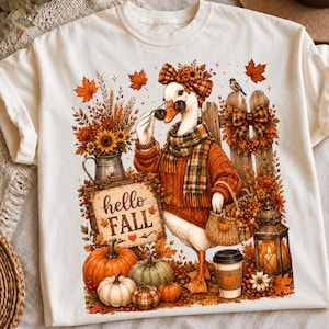 May include: A cream-colored t-shirt featuring a graphic of a goose wearing a plaid scarf, headband, and sunglasses. The goose holds a woven basket near a sign that says hello FALL, pumpkins, a lantern, and autumn foliage.