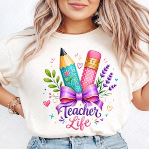 May include: A cream-colored t-shirt featuring a graphic print of a pencil and a pink eraser tied together with a purple bow. The design includes green leaves, purple lavender sprigs, small hearts, and the text Teacher Life in purple script.
