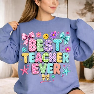 May include: A periwinkle blue crewneck sweatshirt featuring a colorful graphic print that reads BEST TEACHER EVER in bubble letters. The design includes pink and yellow checkered patterns, a smiley face, flowers, bows, and starfish icons.
