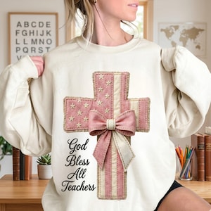 Op de afbeelding: Een crèmekleurige sweater met ronde hals en een grote afbeelding van een kruis in een roze en wit Amerikaans vlagpatroon. Een roze strik is in het midden van het kruis geplaatst en de tekst "God Bless All Teachers" staat in zwarte letters.
