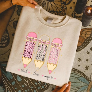May include: A beige crewneck sweatshirt featuring a graphic of three pencils with pink erasers and bows. The pencils have leopard print patterns in pink, black, and light pink. Below the pencils, the text reads "teach love inspire" with small pink hearts.