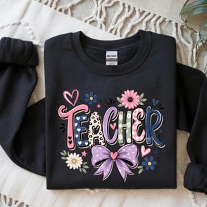 May include: A black crewneck sweatshirt featuring the word TEACHER in colorful, patterned letters. The design includes a pink heart, a large purple bow, and various floral accents in shades of pink, blue, and white, with leopard print details.