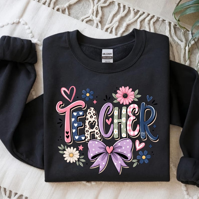 May include: A black crewneck sweatshirt featuring the word TEACHER in colorful, patterned letters. The design includes a pink heart, a large purple bow, and various floral accents in shades of pink, blue, and white, with leopard print details.