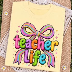 May include: A pale yellow t-shirt featuring a graphic print of a large, multi-colored rainbow bow above the text "teacher life" in a bubbly, textured font. The design includes small star shapes and colorful sprinkles scattered around.