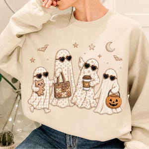 May include: A beige crewneck sweatshirt featuring a graphic of four ghosts wearing heart-shaped sunglasses. The ghosts are decorated with various patterns and are holding a small cat, a tote bag, a coffee cup, and a jack-o'-lantern bucket.