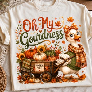 May include: A cream-colored t-shirt featuring a graphic of a duck wearing a plaid scarf, headband, and glasses, pulling a wooden wagon filled with pumpkins and sunflowers. The text reads Oh My Gourdness and Pumpkin Patch, surrounded by autumn leaves.