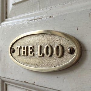 Könnte beinhalten: Ein ovales Messingschild mit der Aufschrift "THE LOO" ist an einer verwitterten, cremefarbenen Holztür angebracht. Das Schild ist mit Schrauben befestigt. Ein Schlüssel hängt im Schloss, und eine Toilette ist im Hintergrund zu sehen.
