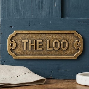 Könnte beinhalten: Rechteckige Messingplakette mit den Worten "THE LOO" in erhabenen Großbuchstaben. Die Plakette hat dekorative Schnörkel an den Rändern und ist auf einer blauen Holzoberfläche montiert.