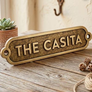 May include: A rectangular, bronze-colored sign with the words "THE CASITA" in raised lettering. The sign has rounded edges and holes for mounting. A small succulent in a terracotta pot sits to the left. The sign rests on a wooden surface.