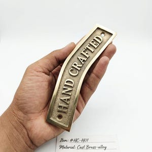 Könnte beinhalten: Eine gebogene rechteckige Plakette aus Messinglegierung mit dem geprägten Schriftzug "HAND CRAFTED". Die Plakette hat eine gealterte Messingoberfläche mit polierten Kanten. Der Artikel ist mit #HC-H01 gekennzeichnet und stammt von Artisanal Forge.