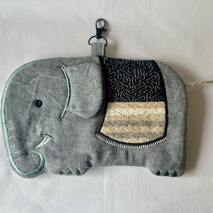May include: A gray fabric elephant-shaped pouch featuring a black metal lobster clasp at the top. The elephant has a black eye, a white tusk, and a decorative saddle patch with a black swirling pattern above horizontal tan and gray stripes.