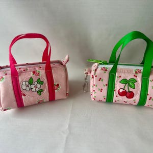 May include: Two small cylindrical fabric bags with pink floral patterns. The left bag has bright pink handles and a white flower patch. The right bag features green handles, a green zipper, and a red cherry patch. Both have metal clips.