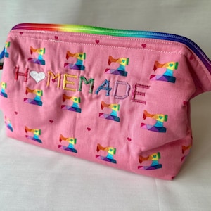 May include: A pink zippered pouch featuring a repeating pattern of rainbow-colored sewing machines and small hearts. The word HOMEMADE is embroidered on the front in multi-colored thread, with a white heart replacing the letter O.