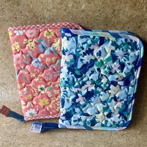 May include: Two rectangular quilted fabric cases with zipper closures. One case features a coral background with a multi-colored floral print and a gingham trim. The other case has a light blue background with a dark blue and green floral pattern.