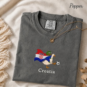May include: A charcoal gray t-shirt featuring a graphic of a green frog character riding a white goose. The frog wears a cape with the Croatian flag design. A soccer ball is near the goose's foot. The word Croatia is printed below the graphic.