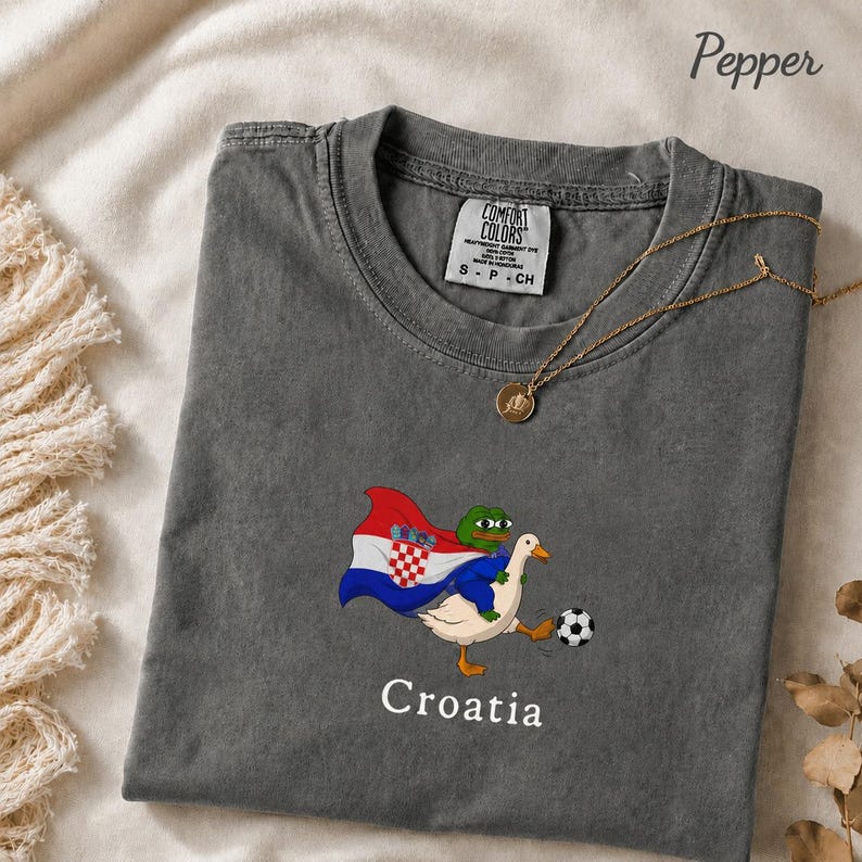 May include: A charcoal gray t-shirt featuring a graphic of a green frog character riding a white goose. The frog wears a cape with the Croatian flag design. A soccer ball is near the goose's foot. The word Croatia is printed below the graphic.