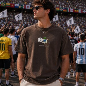 May include: A dark brown short-sleeve t-shirt featuring a graphic of a green frog character riding a white goose, holding a white flag with a question mark, and a soccer ball. The text "country name" is printed below the graphic.
