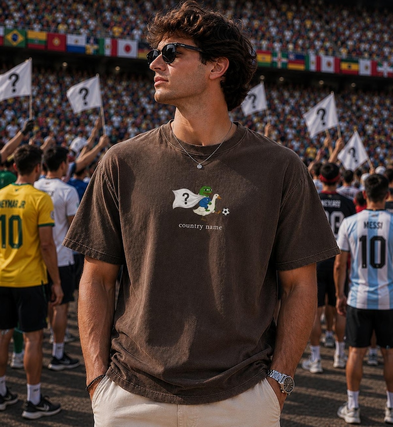May include: A dark brown short-sleeve t-shirt featuring a graphic of a green frog character riding a white goose, holding a white flag with a question mark, and a soccer ball. The text "country name" is printed below the graphic.