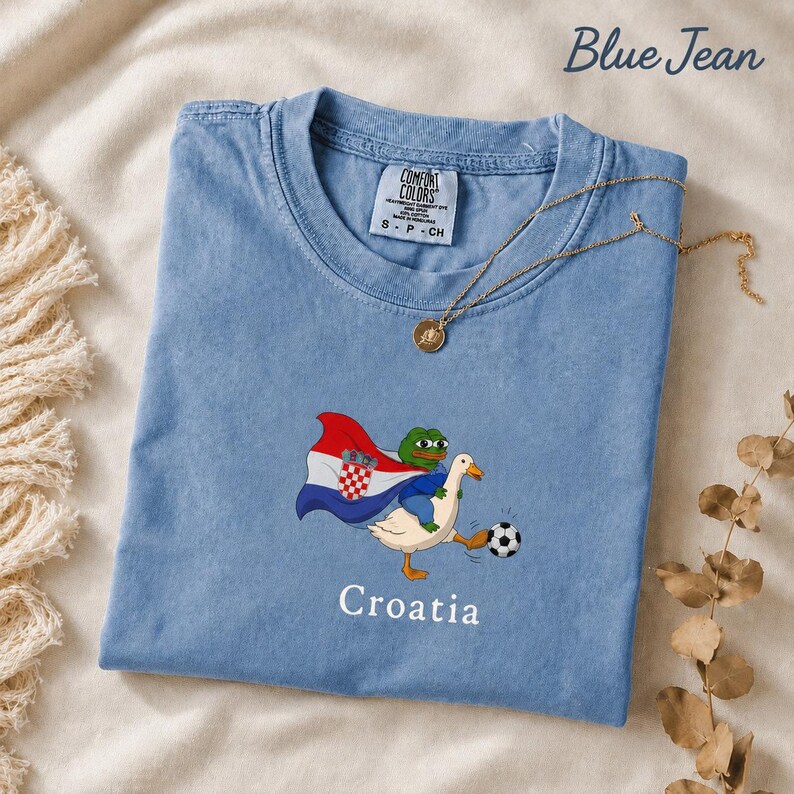 May include: A blue jean colored t-shirt featuring a graphic of a green frog riding a white duck. The duck is kicking a soccer ball, and the frog is wearing a cape with the Croatian flag design. The word Croatia is printed below the graphic.