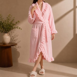 May include: A long, light pink plush robe featuring a shawl collar with ruffled trim, long sleeves with ruffled cuffs, a matching tie belt at the waist, and two front patch pockets. The robe has a soft, fuzzy texture and a ruffled hemline.