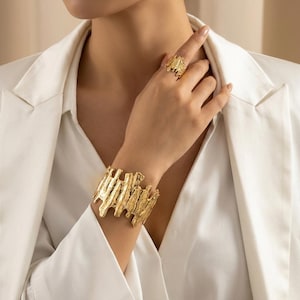 Peut inclure: Un bracelet manchette doré et une bague assortie, tous deux dotés d'une surface texturée, organique et fondue. Le large bracelet manchette entoure le poignet, tandis que la bague repose sur le doigt, présentant un design aux bords irréguliers.