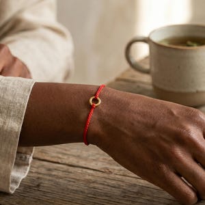 Puede incluir: Una pulsera fina de cordón rojo con un pequeño dije circular de metal dorado centrado en la muñeca. La pulsera consiste en un cordón rojo liso y sencillo atado de forma segura, resaltando el contraste entre la cuerda roja vibrante y el anillo metálico dorado.