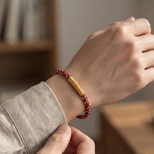 Pode incluir: Uma pulseira trançada com um padrão tecido de fios vermelhos, pretos e dourados. A pulseira inclui uma conta central cilíndrica de metal dourado com uma textura fina e canelada que envolve a estrutura do cordão trançado.