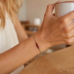 Puede incluir: Una pulsera delicada que presenta un cordón fino trenzado en tonos verdes y amarillos, acentuado con tres pequeñas cuentas redondas de color rojo intenso colocadas centralmente a lo largo de la hebra, creando un accesorio de muñeca sutil.