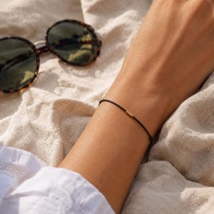 Könnte beinhalten: Ein dünnes schwarzes Kordelarmband mit einer kleinen goldfarbenen zylindrischen Perle. Das Armband wird am Handgelenk getragen und befindet sich neben einer Sonnenbrille mit dunklem Schildpattrahmen und dunklen Gläsern auf einem cremeweißen Stoff.