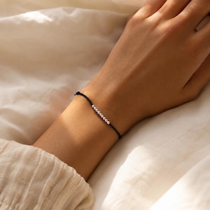 May include: A delicate bracelet featuring a thin black cord adorned with a single row of nine small, round, metallic silver beads centred on the wrist. The minimalist design showcases a smooth, polished finish on the beads against the dark cord.