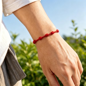 May include: A red knotted bracelet. The bracelet is made of a thin, twisted cord with seven evenly spaced knots. The bracelet is worn on a wrist, with a blurred background of green foliage and a blue sky.