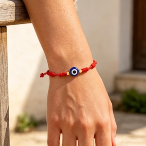 May include: Red string bracelet featuring a blue and white evil eye charm with gold-colored beads. The bracelet is knotted and adjustable, designed for wrist wear. The evil eye is a traditional symbol of protection.