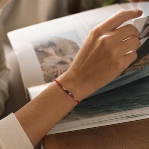 May include: A thin red cord bracelet featuring a small central design composed of two metallic gold-toned cylindrical beads flanking a single black cylindrical bead, secured with a simple knot closure on a slender red string band.