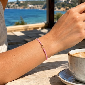 Könnte beinhalten: Ein dünnes, hellrosa geflochtenes Kordelarmband mit einem kleinen, polierten goldfarbenen Metallverschluss. Das Armband wird am Handgelenk getragen und zeichnet sich durch ein schlichtes Design mit einem sicheren, funktionalen Verschluss aus.