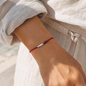 Könnte beinhalten: Ein dünnes rotes Kordelarmband mit einem kleinen silberfarbenen Fisch-Anhänger und zwei winzigen, dunkelroten, kugelförmigen Perlen. Das Armband besteht aus einem einfachen, geflochtenen Schnurmaterial und liegt als zartes Accessoire am Handgelenk.
