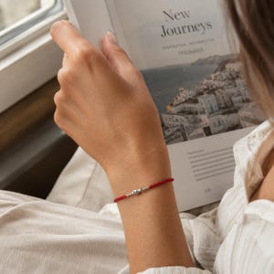 Könnte beinhalten: Ein dünnes rotes Armband aus Kordel mit einem kleinen, silberfarbenen Fisch-Anhänger aus Metall in der Mitte. Das Armband wird am Handgelenk getragen und zeichnet sich durch ein schlichtes Design mit geflochtener Struktur und glänzendem Metall aus.