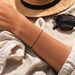 Op de afbeelding: Een fijn zwart koordarmbandje met vijf kleine, gelijkmatig verdeelde goudkleurige kraaltjes. Het armbandje heeft een dunne en minimalistische constructie, ontworpen om nauw om de pols te sluiten met een eenvoudige en ingetogen uitstraling.