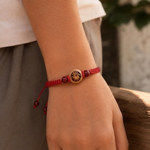 Könnte beinhalten: Rotes geflochtenes Armband mit einem kreisförmigen Holzanhänger mit detailliertem Design. Das Armband enthält rote Perlen und einen Schiebeknotenverschluss. Das Armband wird am Handgelenk getragen.
