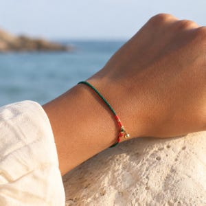 Pode incluir: Uma pulseira delicada com um cordão fino verde-escuro acentuado por uma secção de fio vermelho vibrante, pequenas contas douradas e um único pendente esférico dourado, criando um acessório refinado para o pulso.