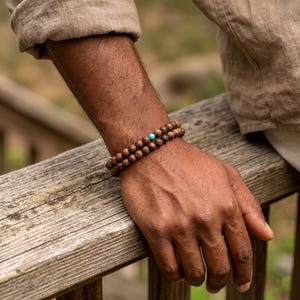 Puede incluir: Una pulsera de cuentas de doble hilo con pequeñas cuentas de madera redondas de color marrón oscuro con una superficie texturizada. Una sola cuenta lisa de color azul turquesa brillante se coloca como punto focal en el diseño.