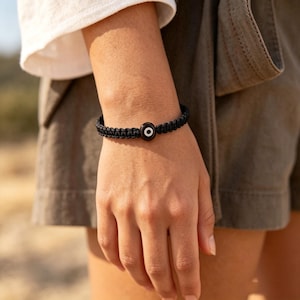 May include: Black braided bracelet featuring a central, round charm with an eye design. The charm has a dark background with a white and black eye detail. The bracelet is worn on a wrist, showcasing the accessory's design and style.