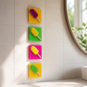 Op de afbeelding: Vier kleurrijke wanddecoraties in de vorm van ijslolly's. Elke ijslolly is gemonteerd op een vierkante tegel. De ijslolly's zijn paars, geel, groen en geel, met houten stokjes. De tegels zijn respectievelijk geel, groen, roze en geel. De tegels zijn gemonteerd op een witte betegelde muur.