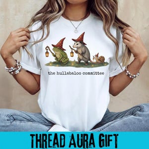 Op de afbeelding: Een wit t-shirt met een afbeelding van een groene kikker, een grijze opossum en een bruine slak, die allemaal puntige rode hoeden met gouden sterren dragen. Elk dier draagt een lantaarntje. De tekst "the hullabaloo committee" staat eronder gedrukt.