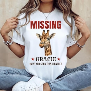 May include: A white short-sleeve t-shirt featuring a graphic of a giraffe head in the centre. Above the giraffe is the red text MISSING. Below the giraffe is the text GRACIE flanked by two red stars, followed by the text HAVE YOU SEEN THIS GIRAFFE?
