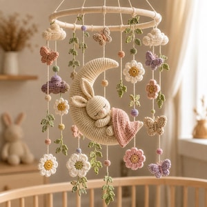 Puede incluir: Un móvil de ganchillo para bebé con una luna creciente de color crema en el centro y un conejito durmiendo bajo una manta rosa. Colgando de un marco circular hay varios elementos de ganchillo que incluyen mariposas, flores blancas y rosas, enredaderas verdes y nubes.
